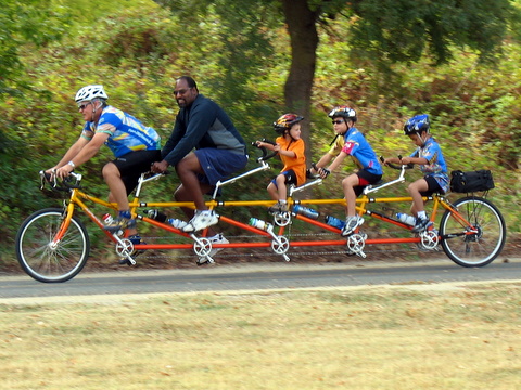 a five-seater bicycle being pedaled by 2 adults and 3 children
